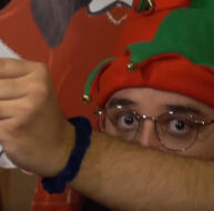a cropped photo of scorpy, a medium-skinned man. he's reaching off camera and his arm is covering his face, so you can only see his christmas elf hat and wide, frightened eyes.